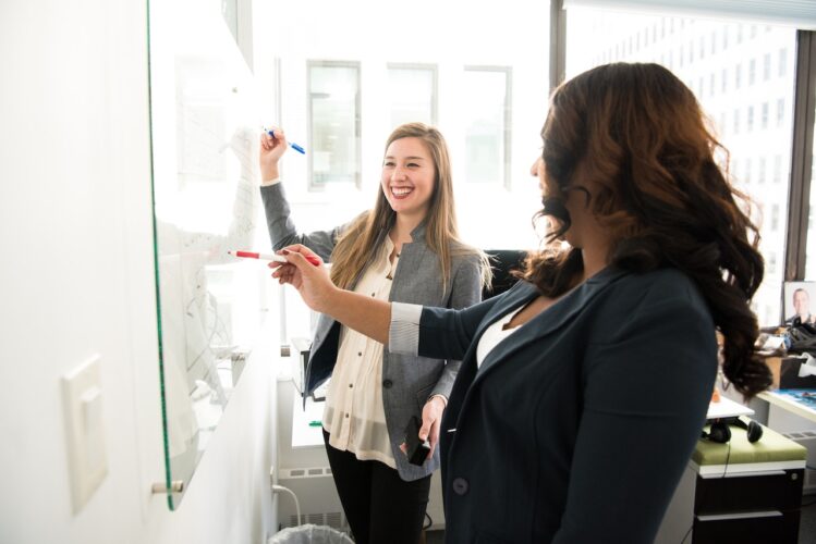 two woman talking in front of dry erase board